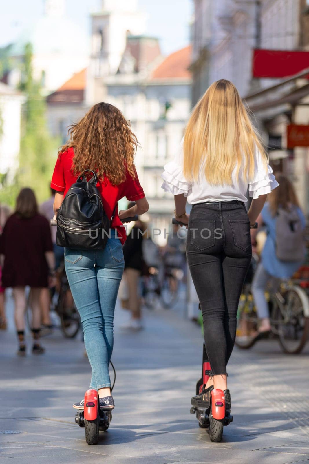 Rear view of trendy fashinable teenager girls riding public rental electric scooters in urban city environment. New eco-friendly modern public city transport in Ljubljana, Slovenia.