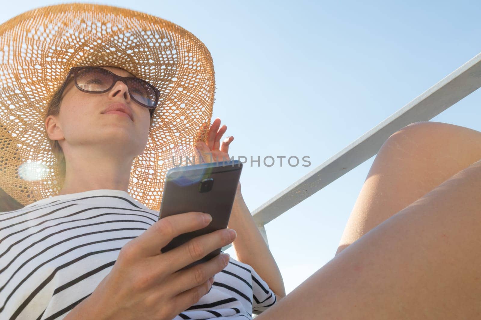 Beautiful, romantic blonde woman on summer vacations traveling by cruse ship ferry boat holding and using mobile phone. Summer vacation lifestyle.