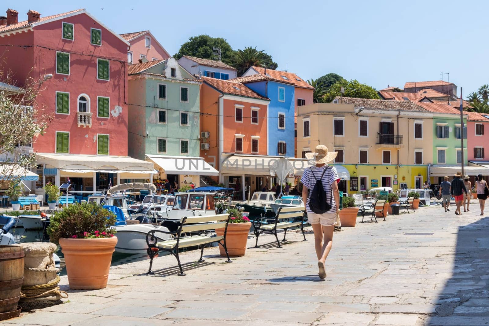 Rear view of beautiful blonde young female traveler wearing straw sun hat sightseeing and enjoying summer vacation in an old traditional costal town of Veli Losinj, Adriatic cost, Croatia.