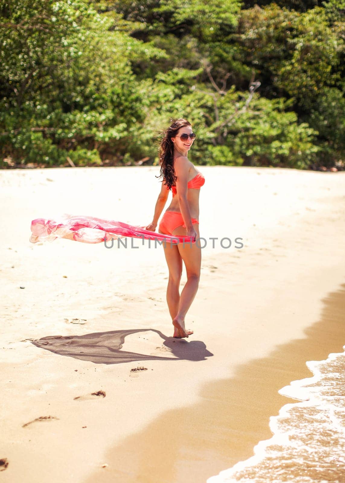Young woman with long dark hair in orange bikini on the beach holding pink scarf in the wind looking back, walking on sandy beach. Nui Bay, Koh Lanta, Thailand
