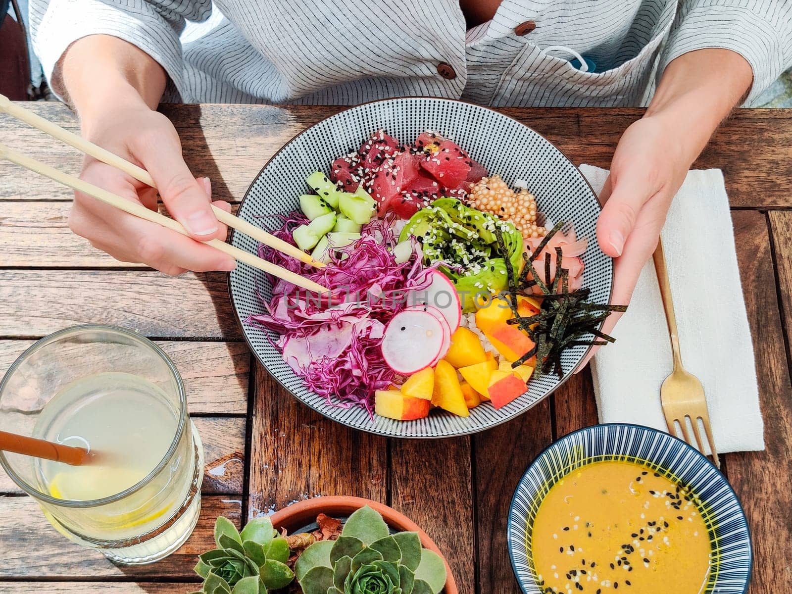 Woman eating tasty colorful healthy natural organic vegetarian Hawaiian poke bowl using asian chopsticks on rustic wooden table. Healthy natural organic eating concept.