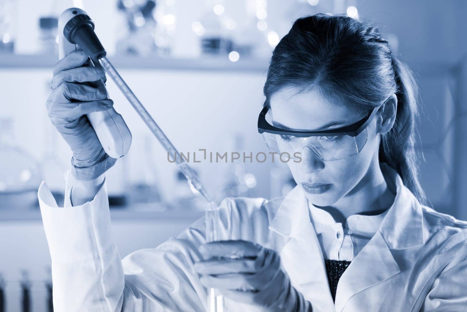 Life scientists researching in laboratory. Focused female life science professional pipetting solution into the glass cuvette. Healthcare and biotechnology concept. Blue toned image.