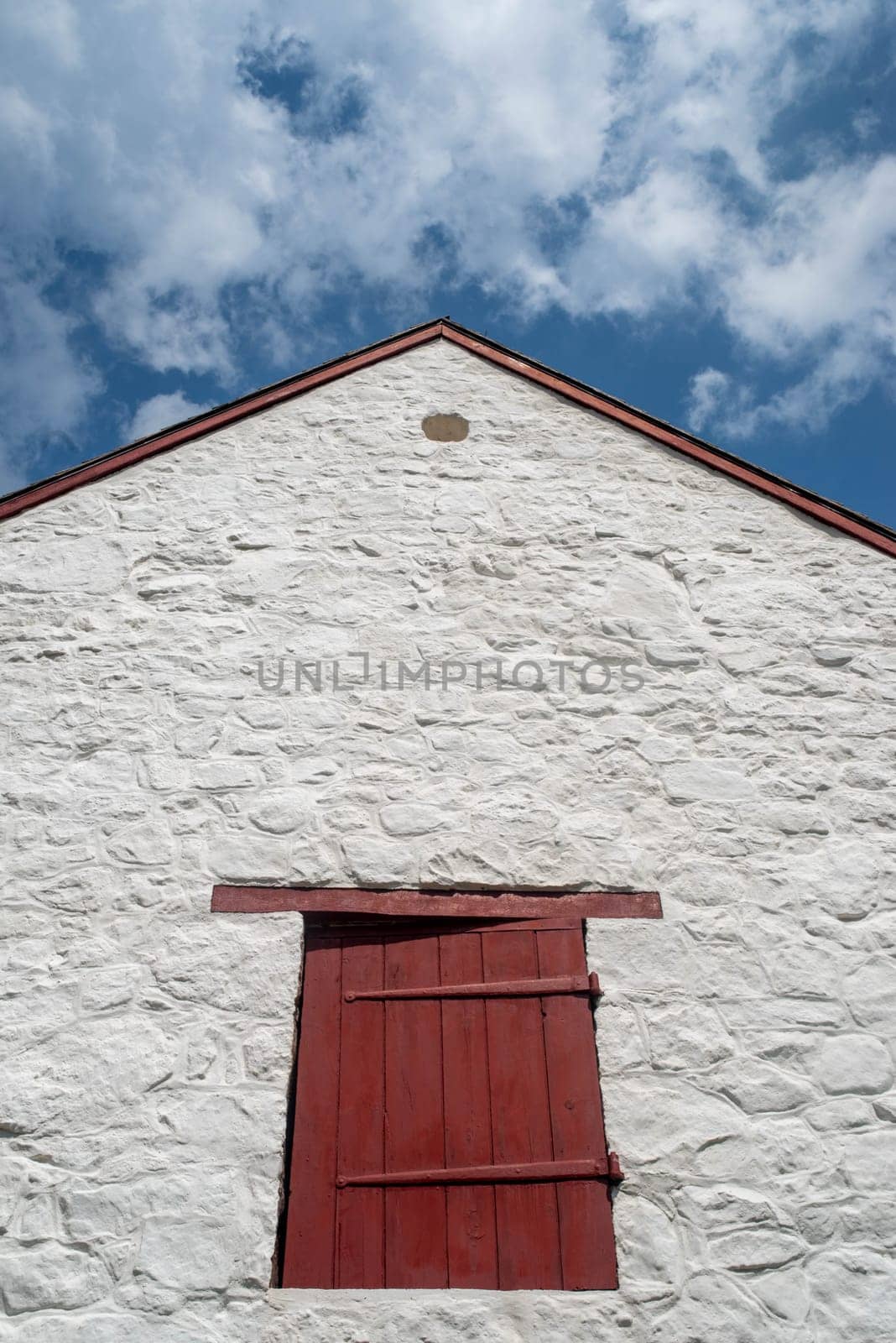 Beautiful whitewashed stone and red painted shutters on a historic colonial American barn at the Hopewell Furnace National Historic Site in Pennsylvania, USA. Close up of peaked roof against blue sky.