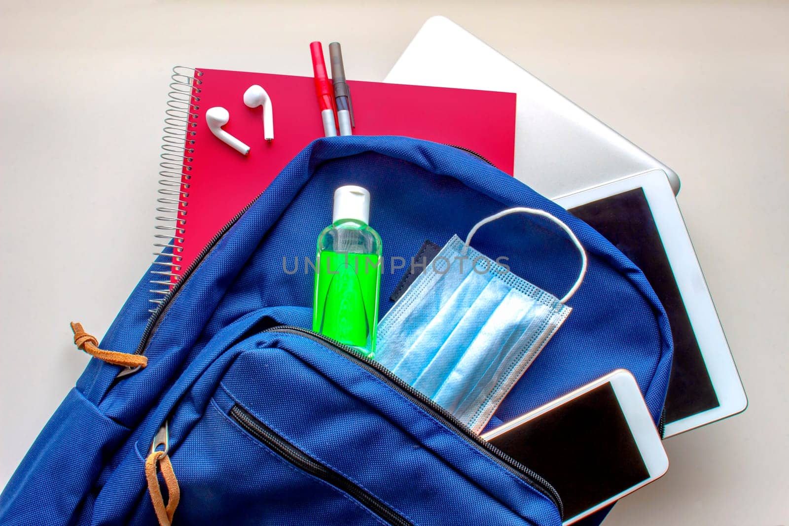 A blue school bag pack with sanitizer and a face mask, pencils earphones, tablet, computer and a smartphone. Back to school during global pandemic. Covid 19.