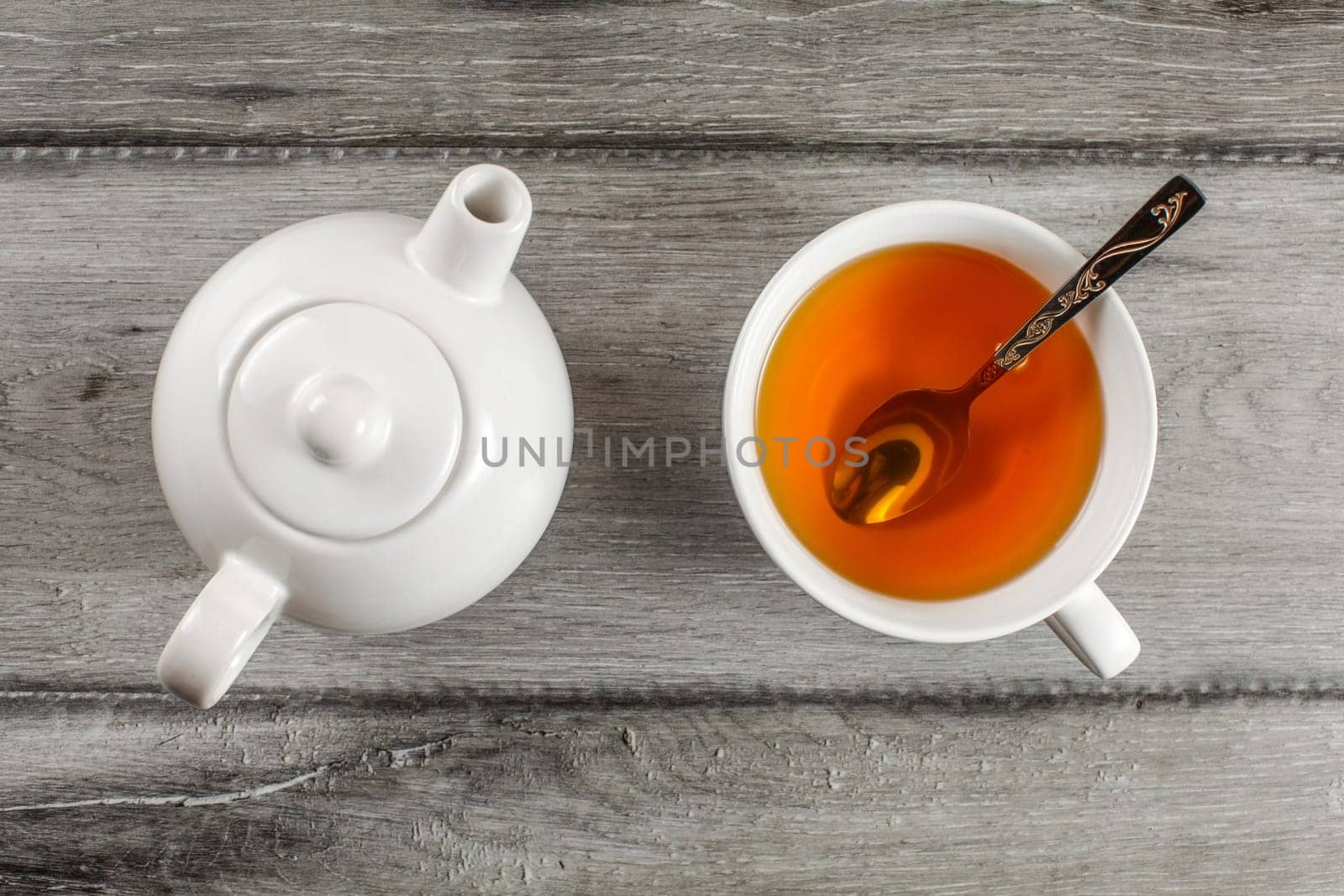 Table top view on small white teapot and cup full of hot amber tea with silver spoon in it, placed on gray wood table.