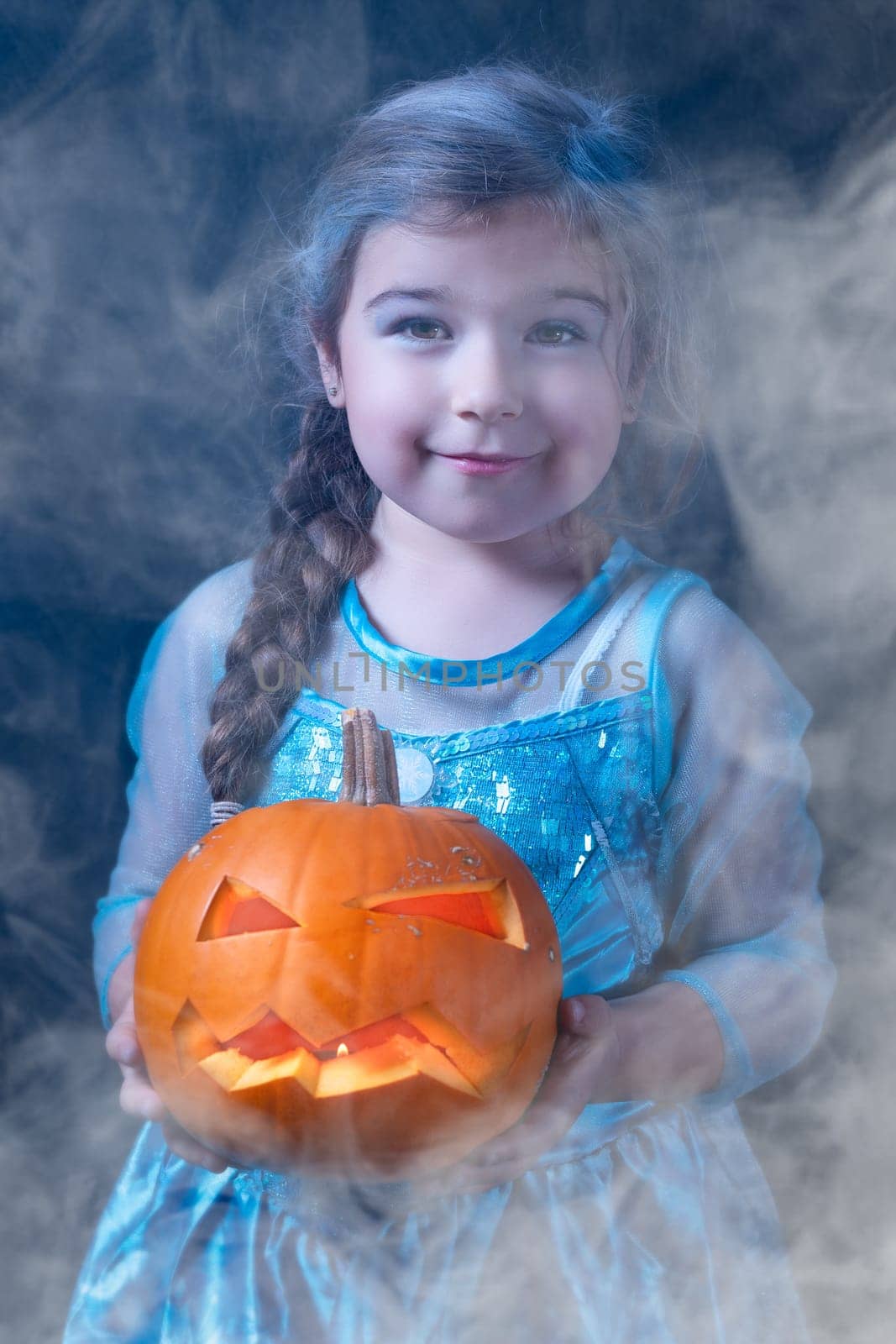 Halloween portrait of young girl stylised for frozen princess in a blue dress. Smiling child holding orange curved pumpkin surrounded by smoke or mist
