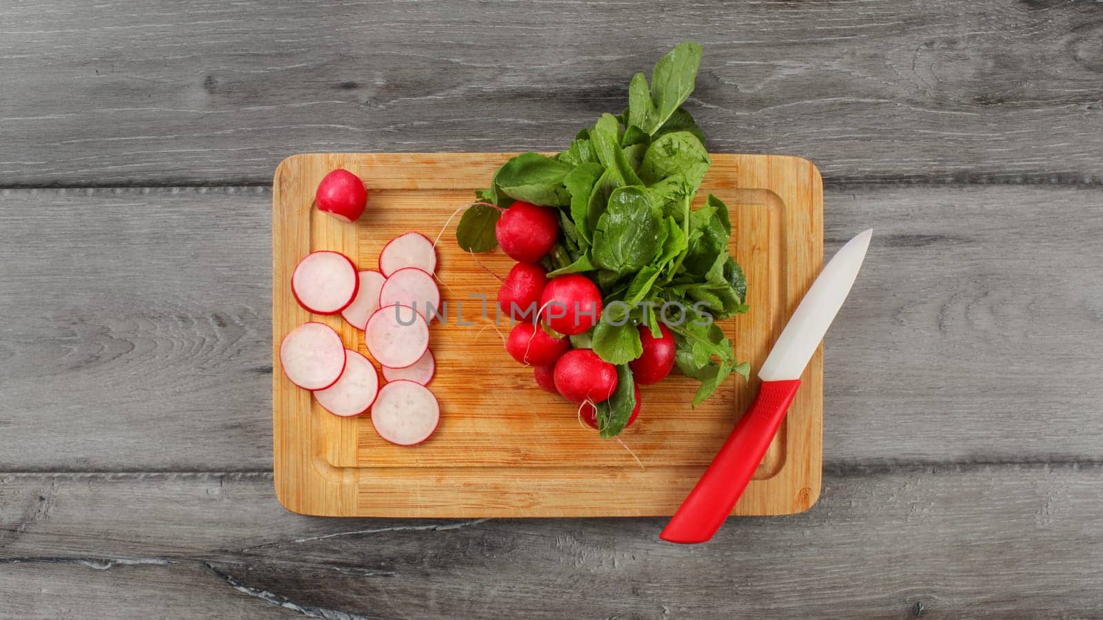 Table top view on old wooden chop board with bunch of bright fresh radish with green leaves, some of them cut in circle pieces with ceramic knife.