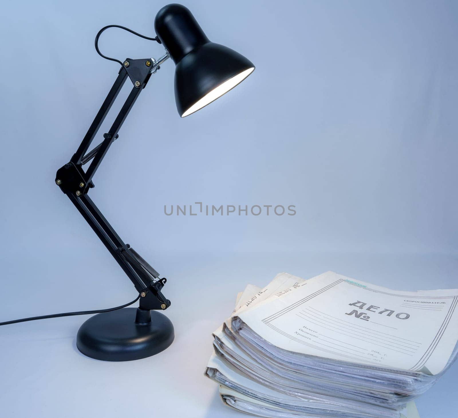 Black adjustable desk lamp casting light on a large stack of documents on the cover. Workspace or office concept with paperwork and study materials