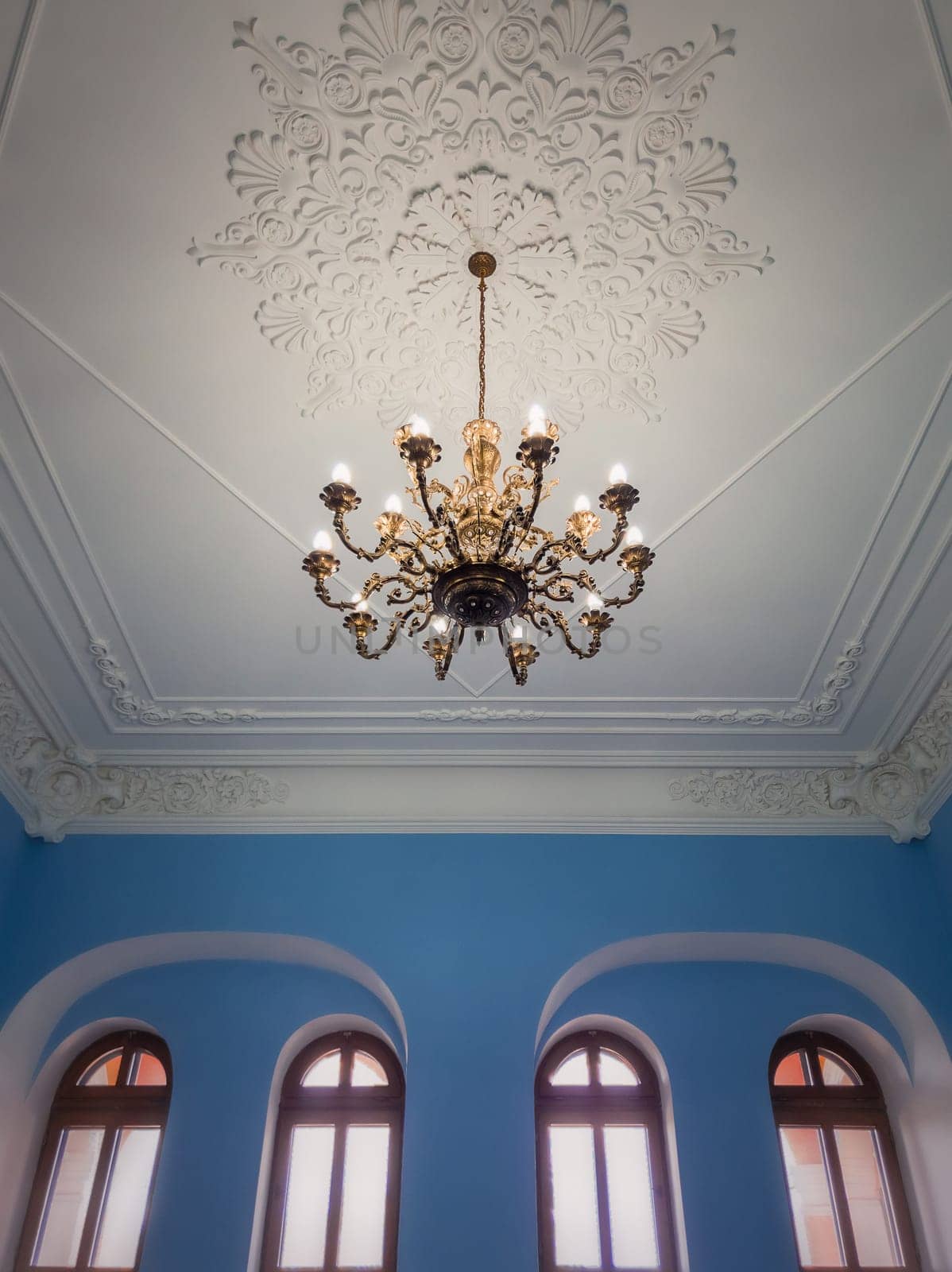 Beautiful golden chandelier suspended down a white ornate ceiling inside a palace room with blue walls and arched windows. Different geometric shapes, ancient interior architecture and design details.