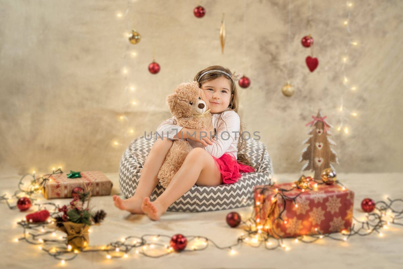 Christmas studio shoot of a cute baby girl that is hugging a teddy bear. Festive, beige background with gifts, Christmas balls and lights
