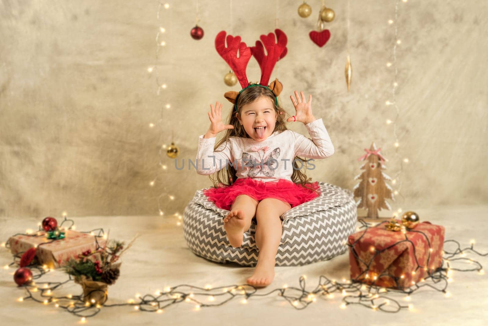Christmas studio shoot of a cute baby girl wearing reindeer headband. Festive, beige background with gifts, Christmas balls and lights