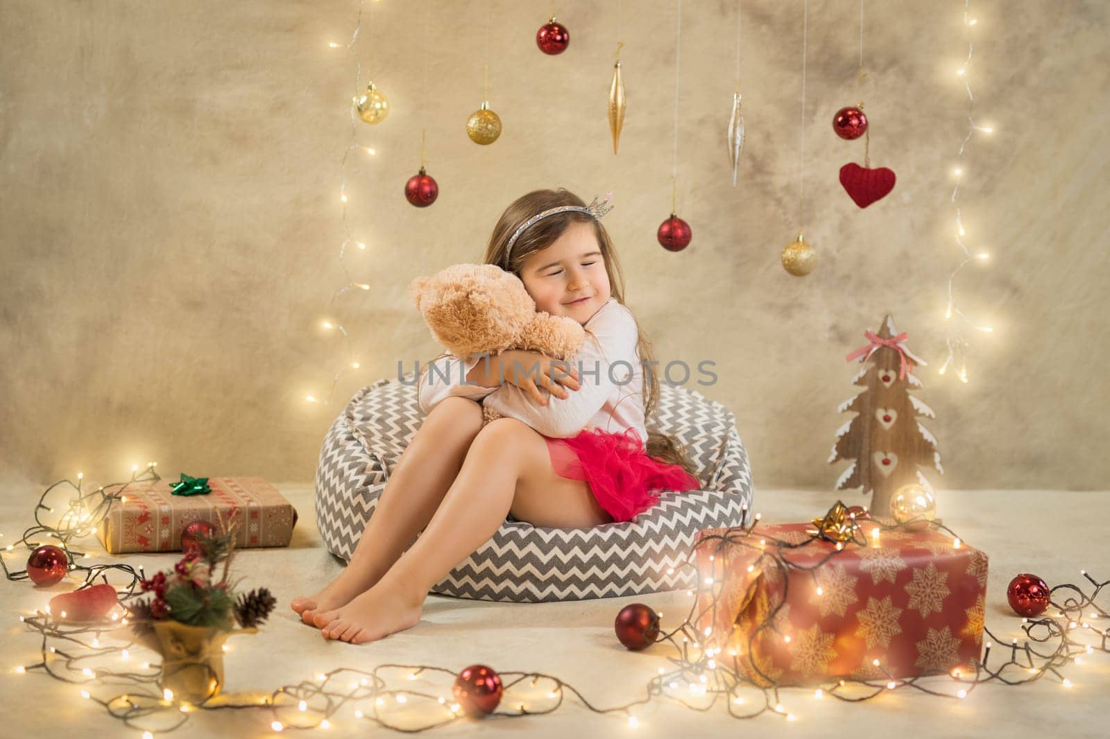 Christmas studio shoot of a cute baby girl that is hugging a teddy bear. Festive, beige background with gifts, Christmas balls and lights