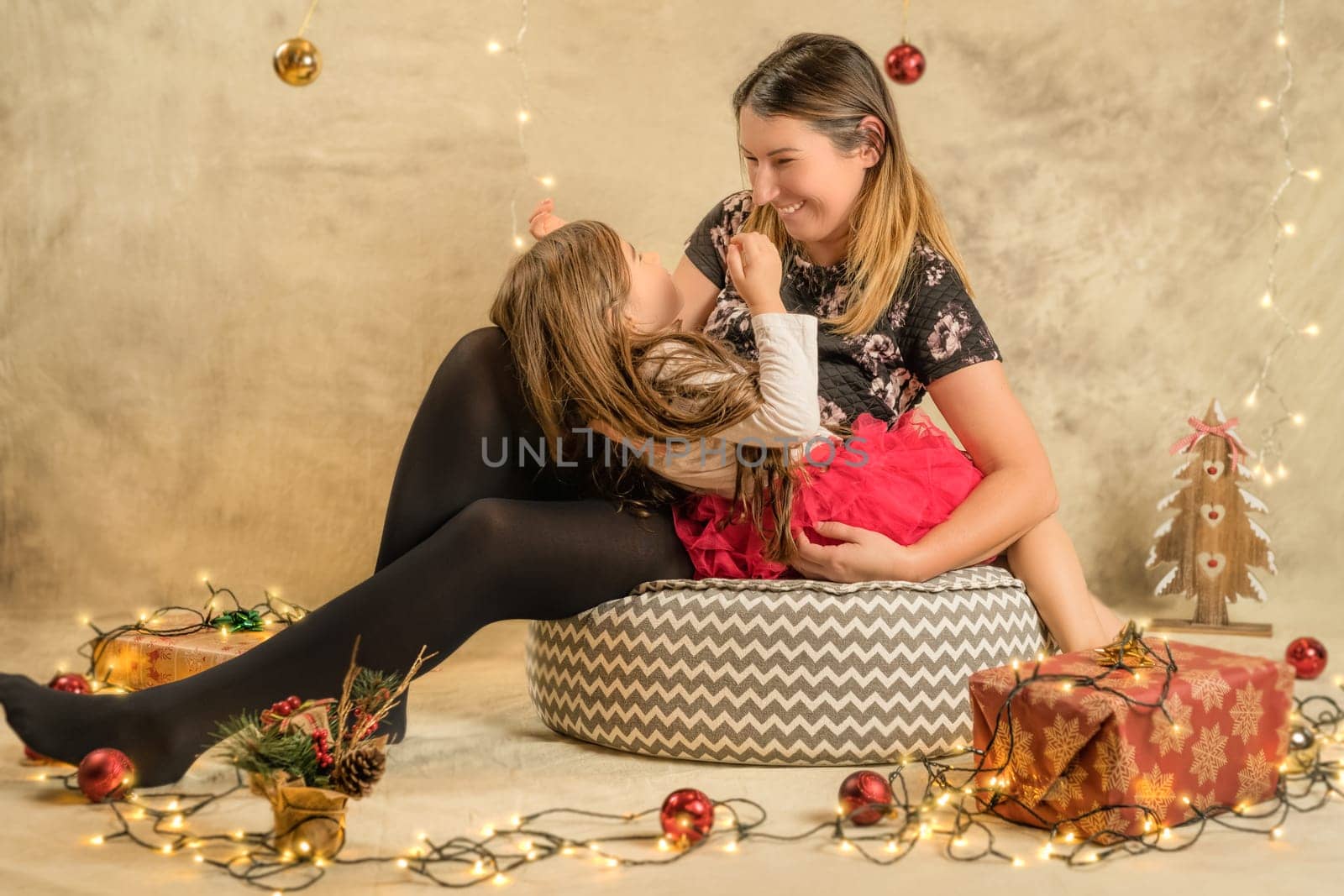 Christmas studio shoot of a cute baby girl and her mother, a happy family. Festive, beige background with gifts, Christmas balls and lights
