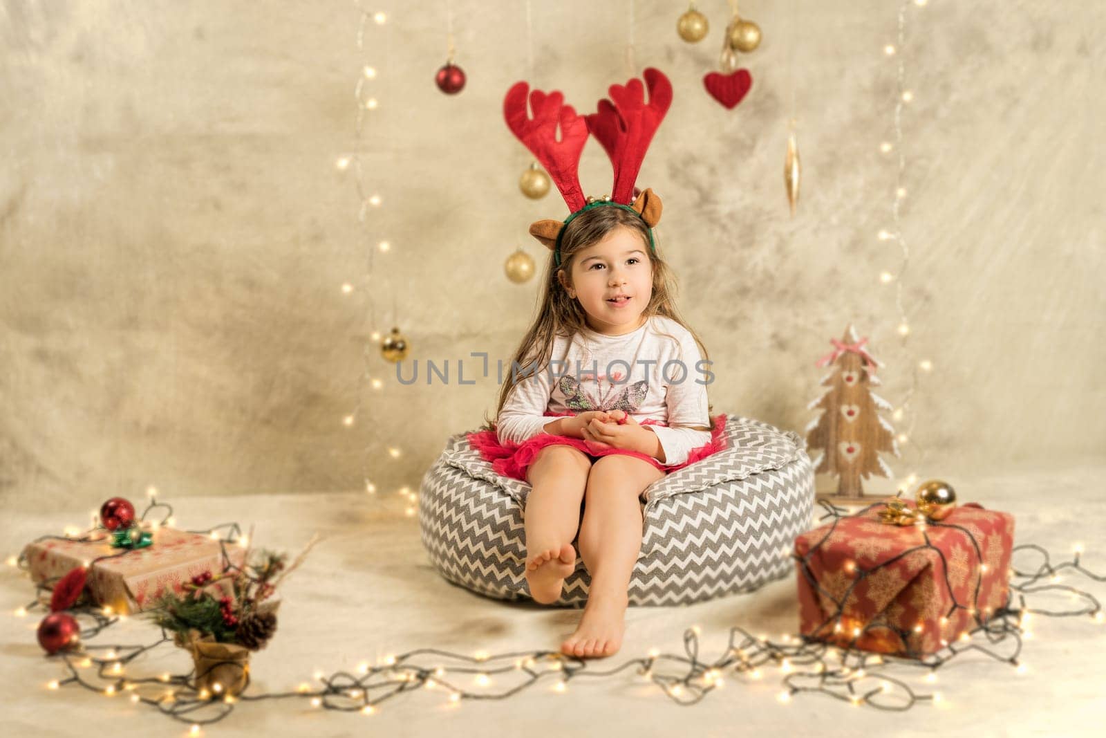Christmas studio shoot of a cute baby girl wearing reindeer headband. Festive, beige background with gifts, Christmas balls and lights