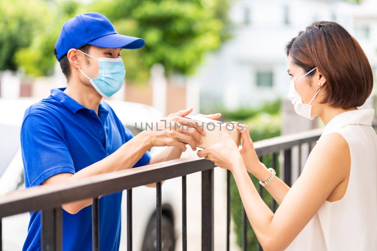 Asian deliver man in blue shirt handling package and give to young woman costumer at house. They wear face mask to reduce coronavirus COVID-19 spreading. Package shopping delivery concept.
