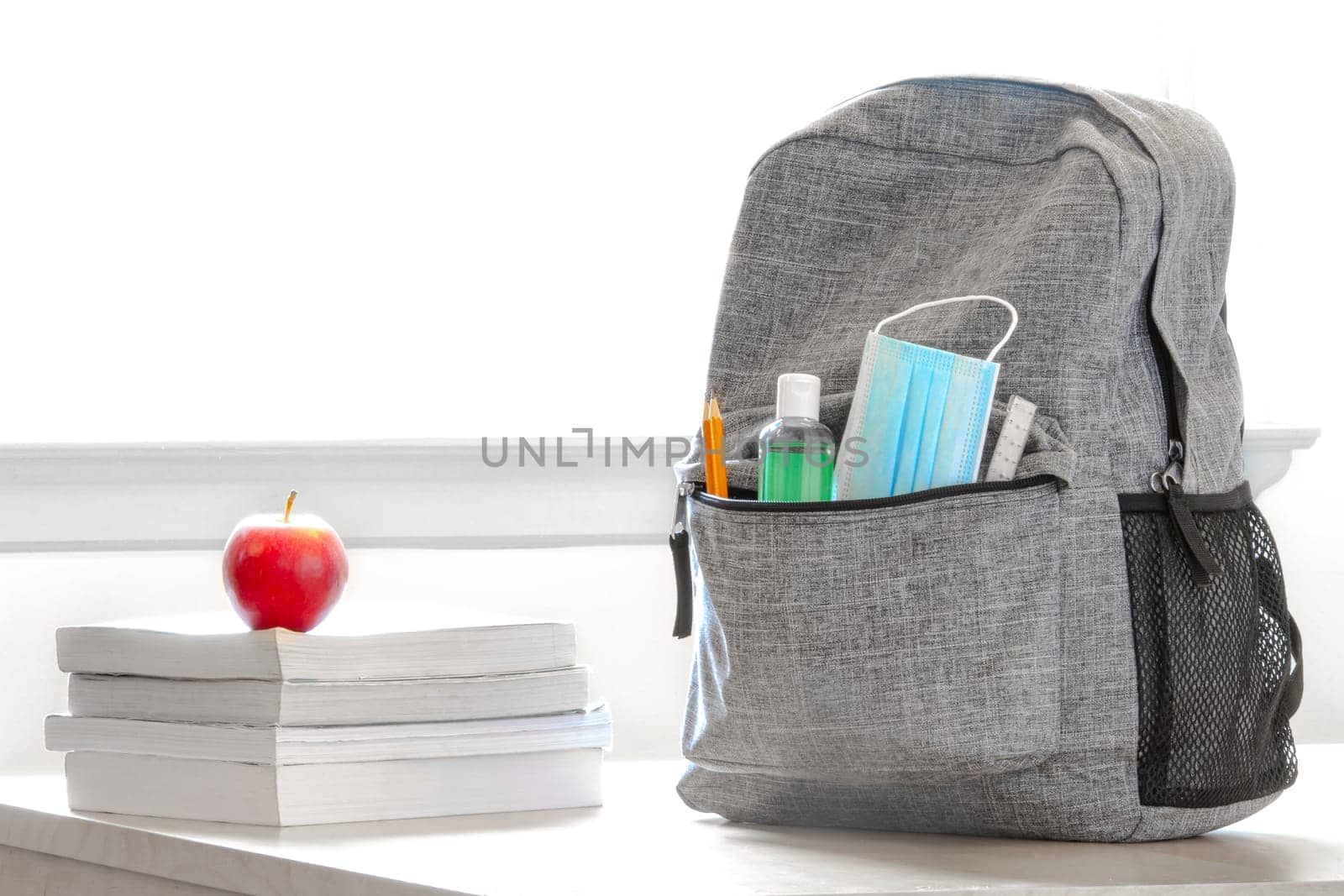 Student school bag pack with sanitizer, a face mask, pencils, books and an apple on a table. Back to school during global pandemic. Covid 19.