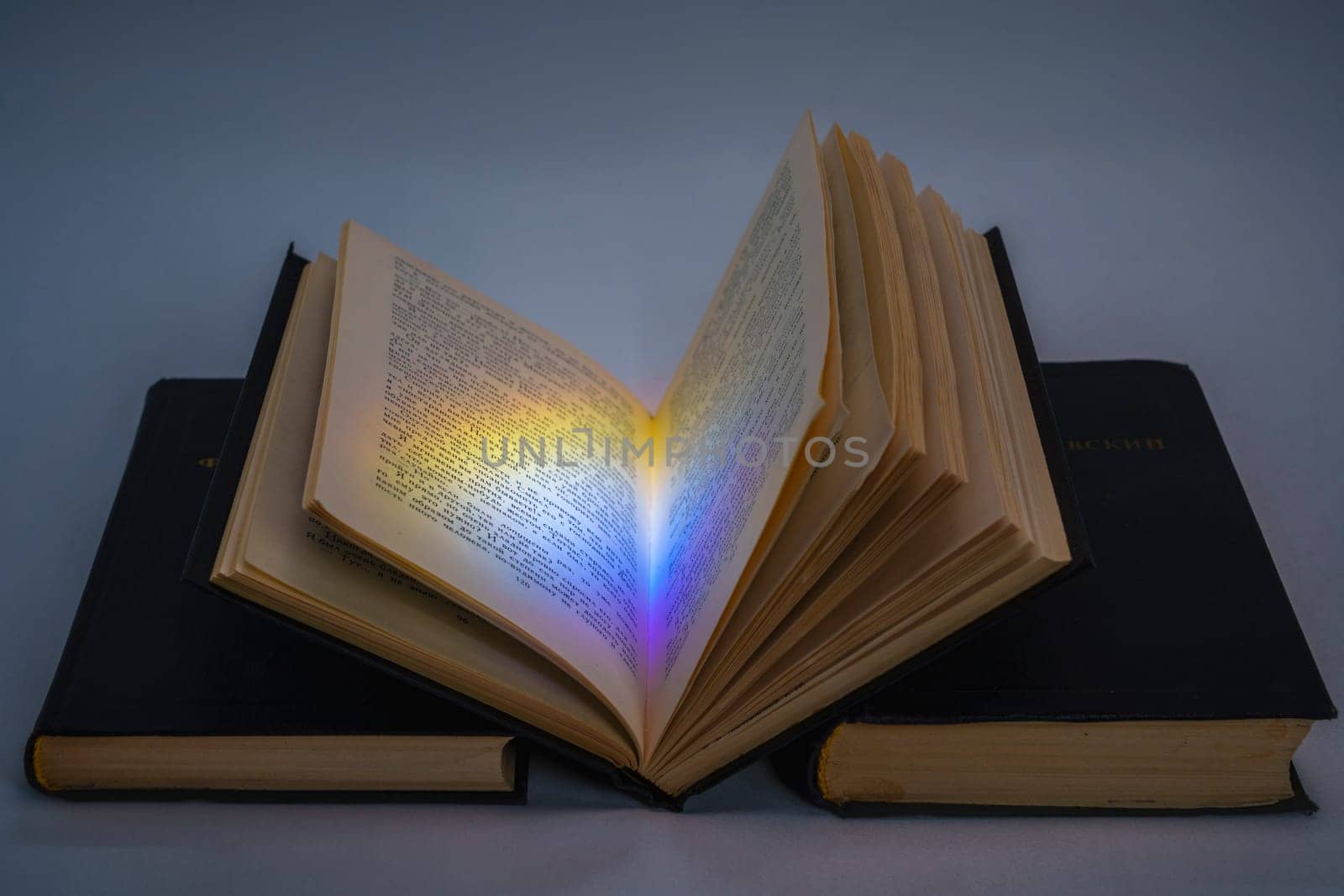 An open vintage book with a colorful light reflection on its pages, resting on two stacked black hardcover books. Symbolic concept of knowledge, mystery, and inspiration