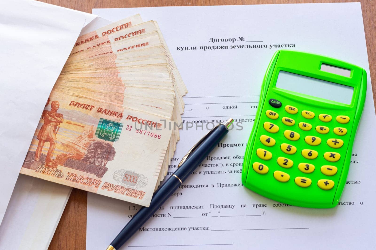 On the table is a contract for the sale of a land plot, a bundle of money in an envelope, a pen and a calculator