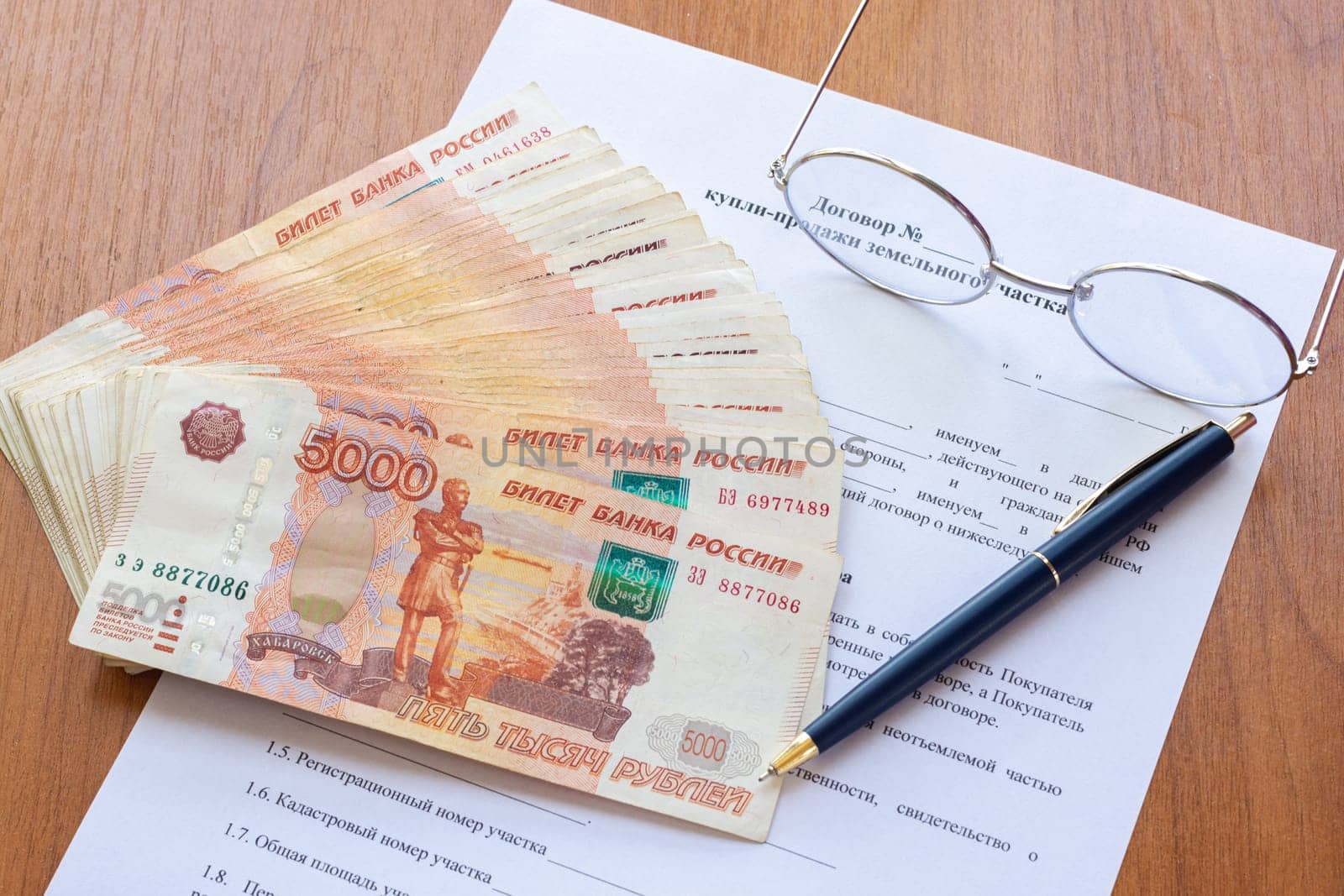 On the table is a contract for the sale of a land plot, a bundle of money, a pen and glasses
