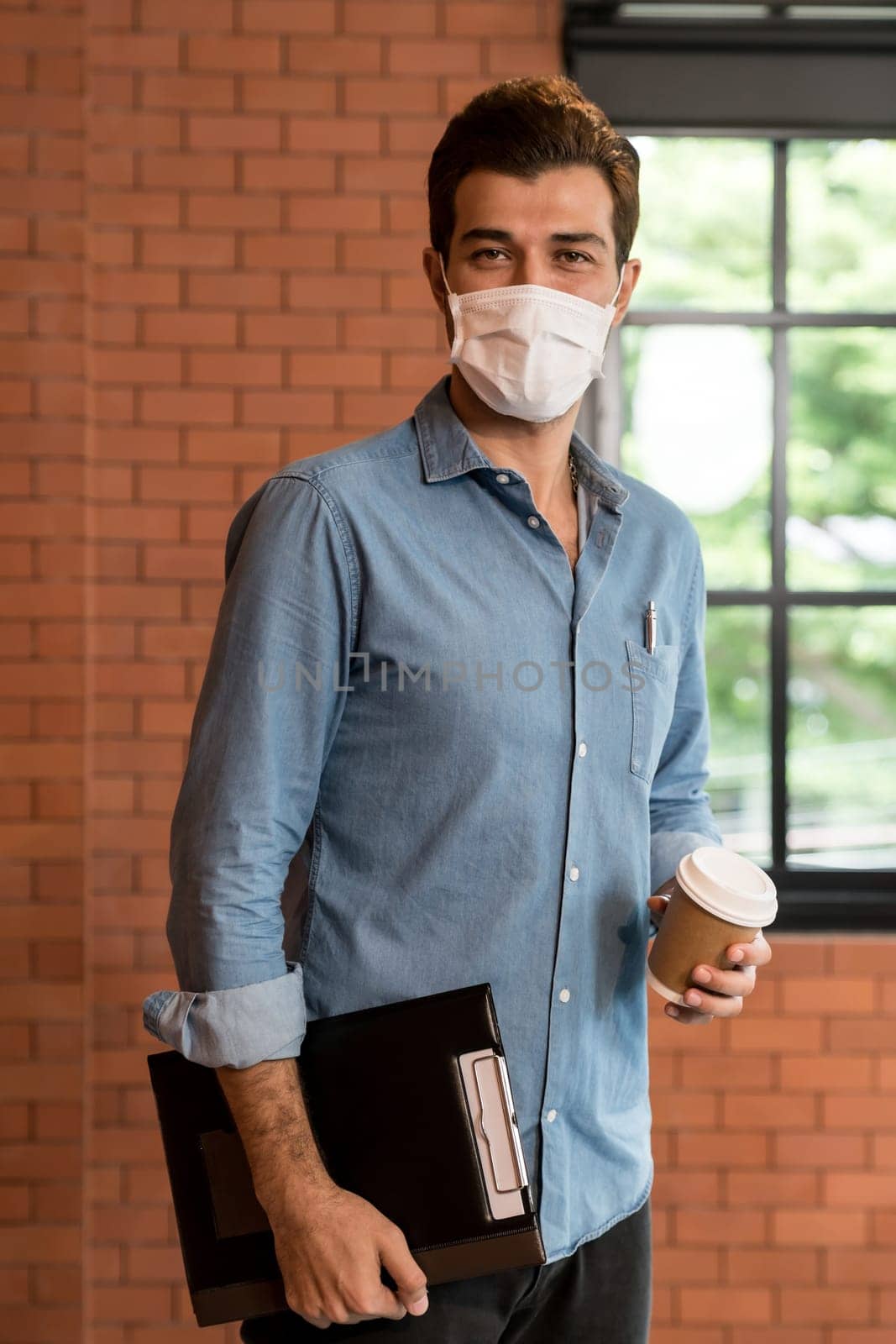 Portrait of middle east caucasian businessman with takeout coffee cup and clipboard in meeting room after lunch break. He wear face mask as new normal office preventing coronavirus COVID-19 spreading.