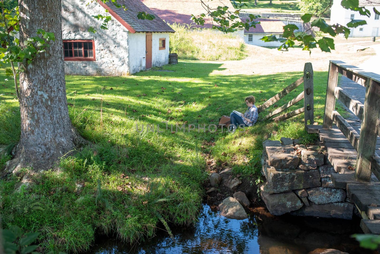 Tranquil early American villiage scene at Hopewell Furnace National Historic Site in Pennsylvania. Young woman relaxes alone in the shade reading a book.