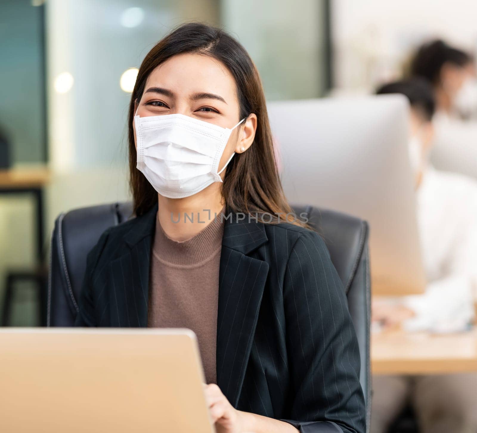 Portrait of asian office employee businesswoman wear protective face mask work in new normal office with interracial colleague in background as social distance practice prevent coronavirus COVID-19.