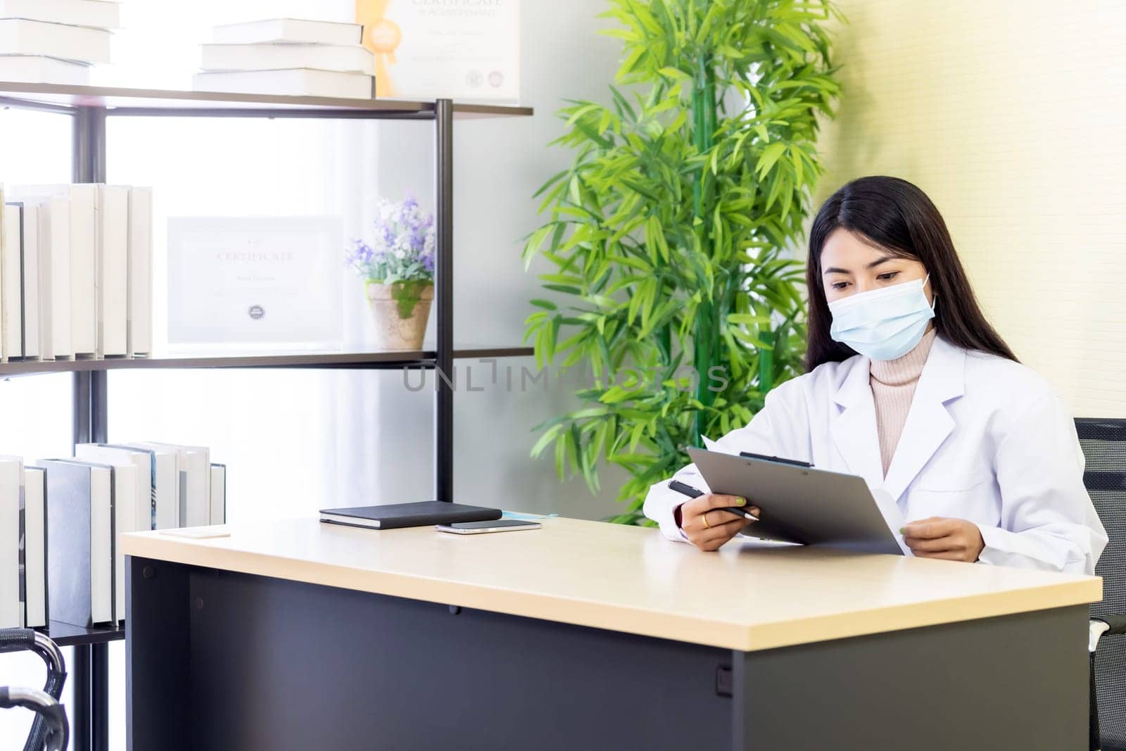 Portrait of asian female doctor wear protective face mask sit in her office room in hospital clinic and reading on patience file before checking patience. New Normal Health care Concept.