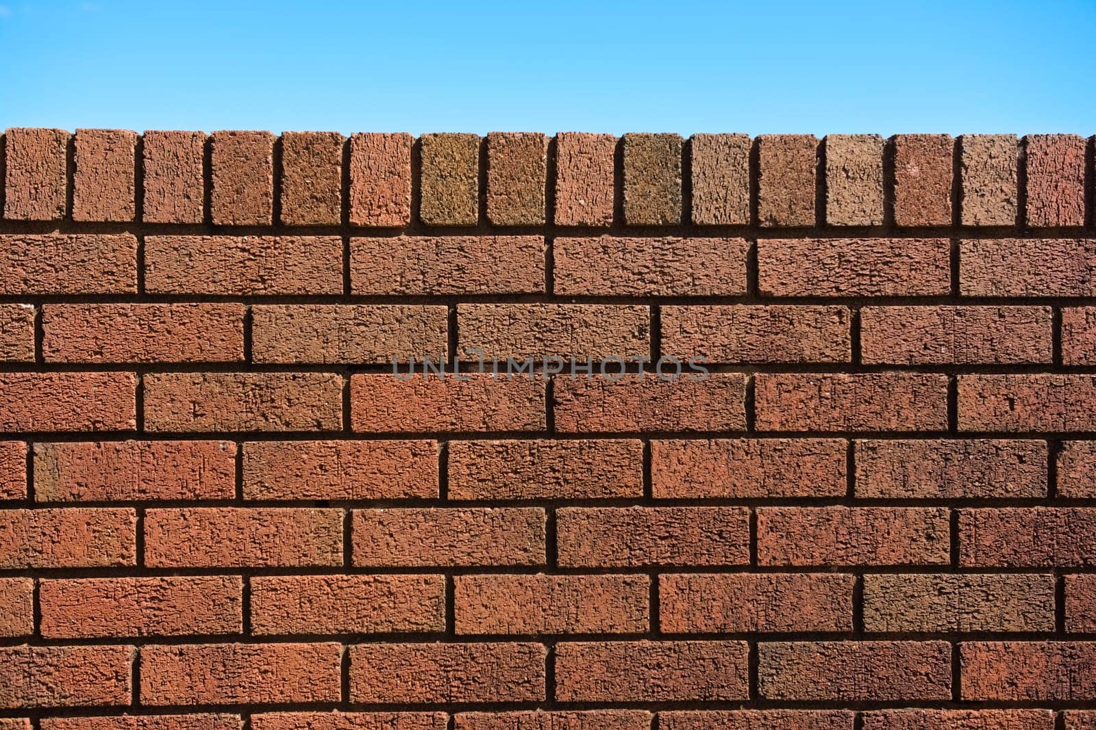 6 - Plain red brick wall background against a bright blue sky background. Sunny summer day. Abstract rough texture with straight dark shadow lines.