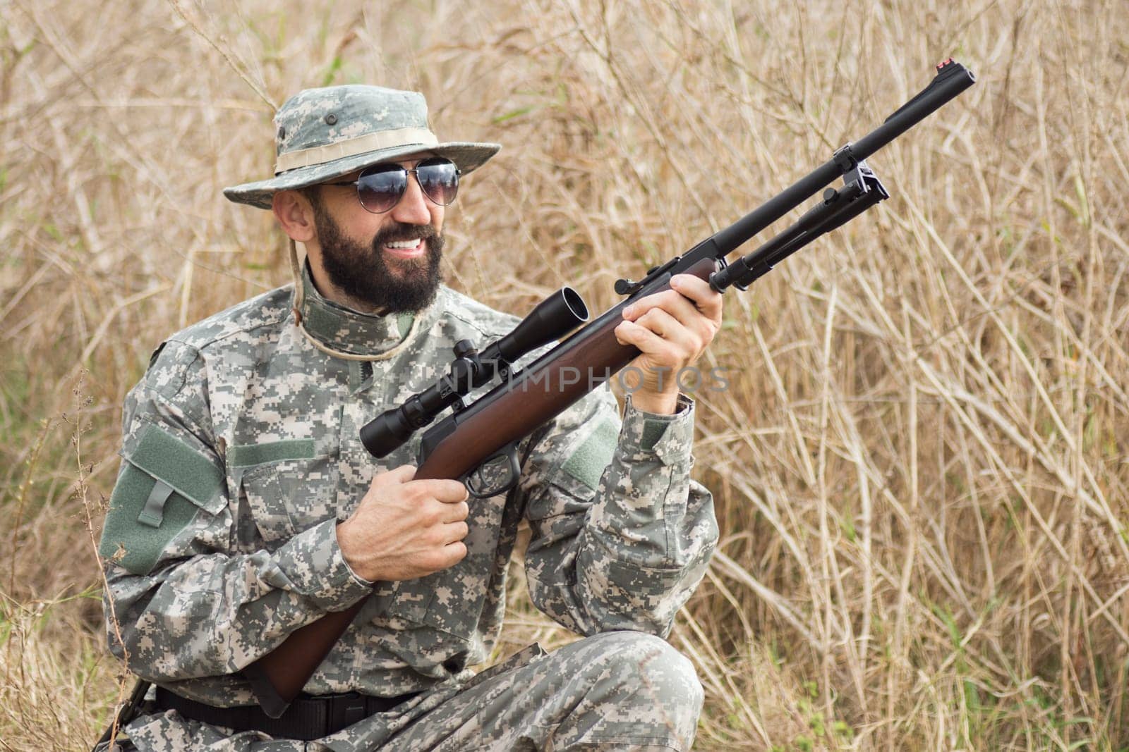 The smiling hunter in military uniform is holding rifle weapon or airgun and squatting in nature.
