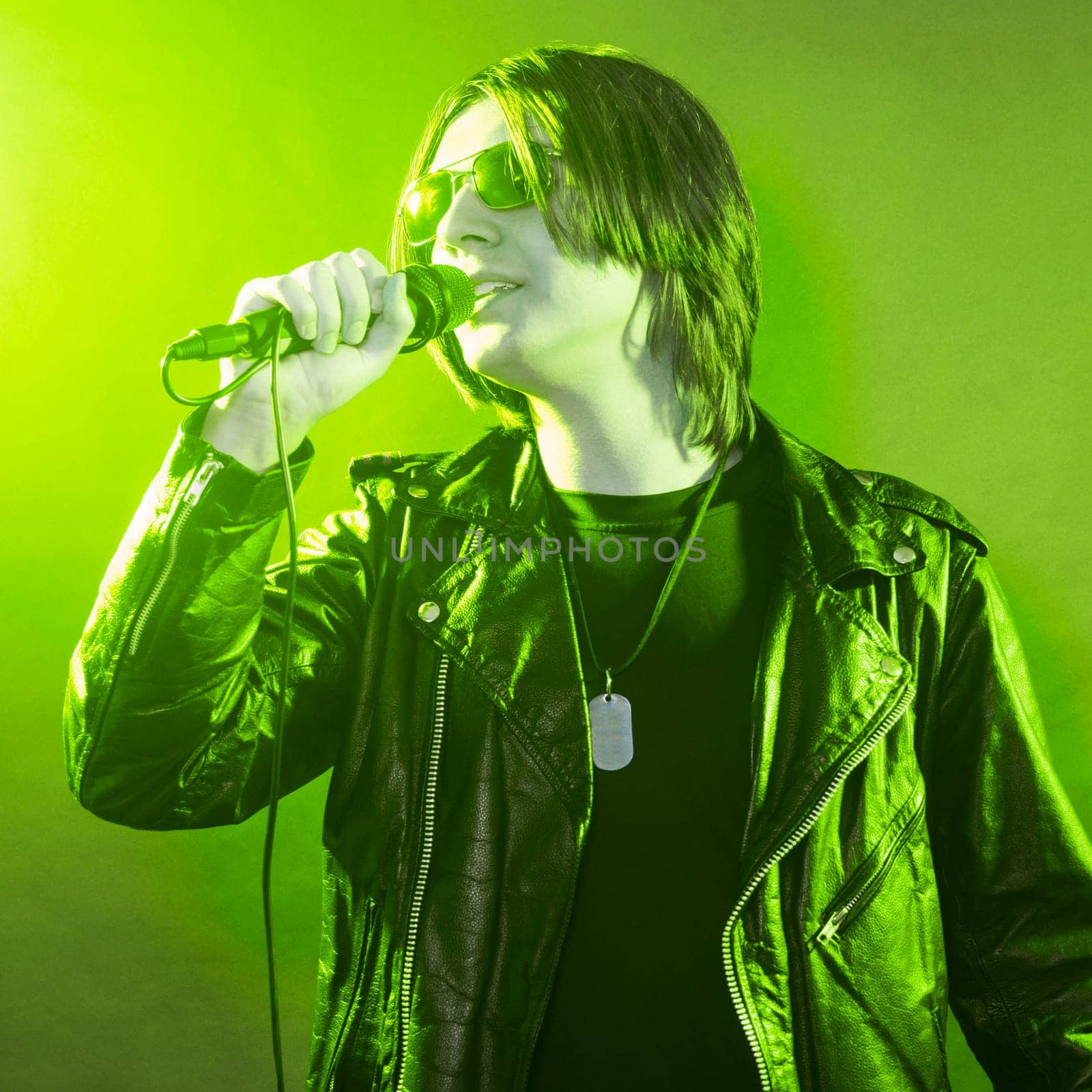 The young guy in black leather jacket is singing rock music on green yellow illuminated stage.