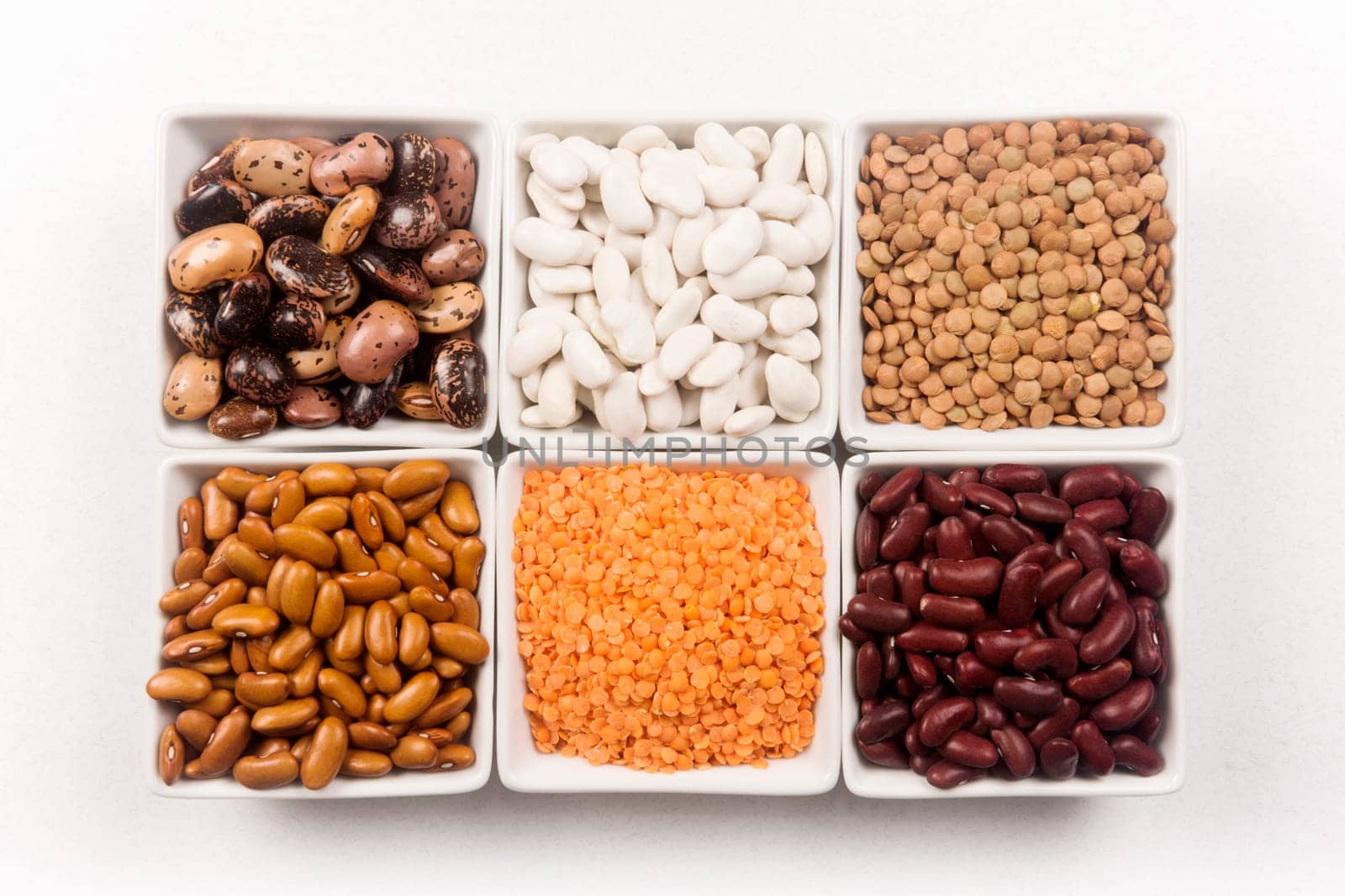 Different legumes seeds as beans and lentils are in white dishes on white background.