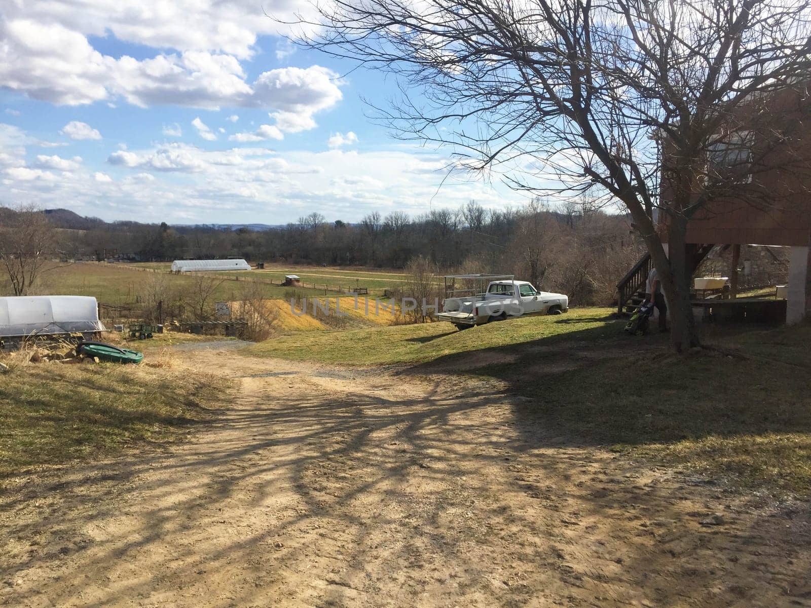 Full frame image of a family farm with greenhouses in the distance and cultivated fileds. Long shadows criss cross the dirt road.