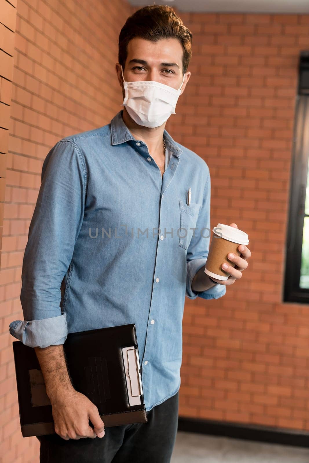 Portrait of middle east caucasian businessman with takeout coffee cup and clipboard in meeting room after lunch break. He wear face mask as new normal office preventing coronavirus COVID-19 spreading.