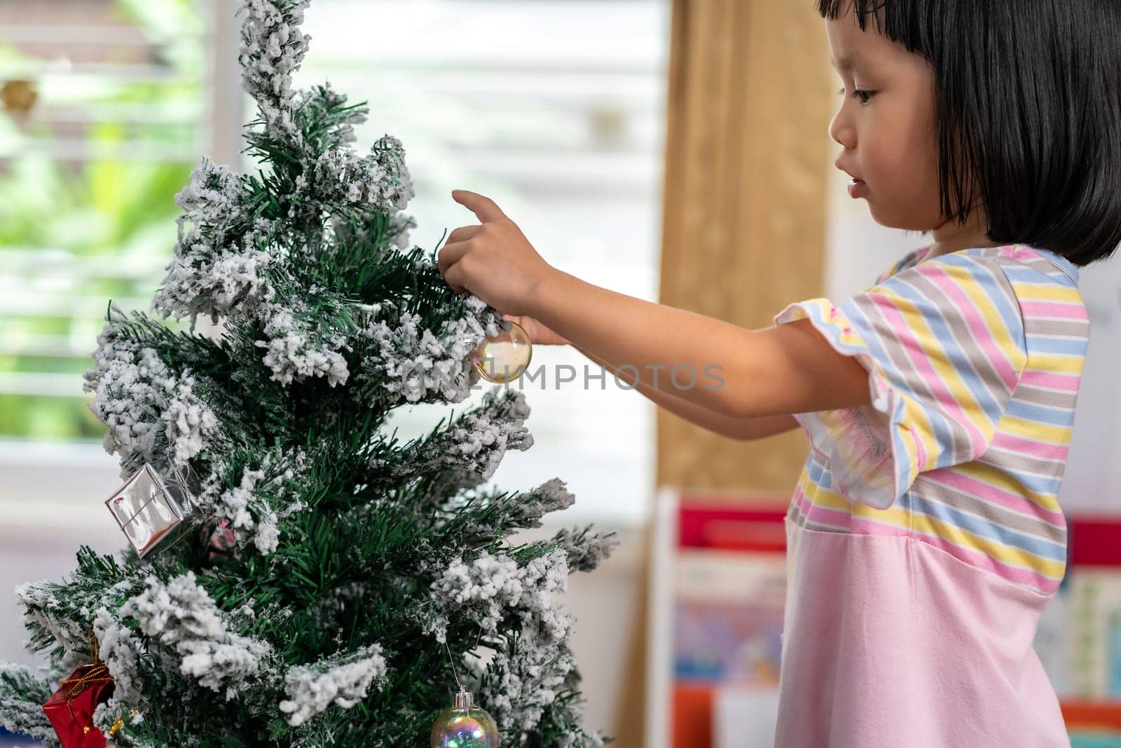Little asian girl decorating a Christmas tree in her living room with christmas ornament preparing for season greeting of Merry Christmas and Happy Holidays.