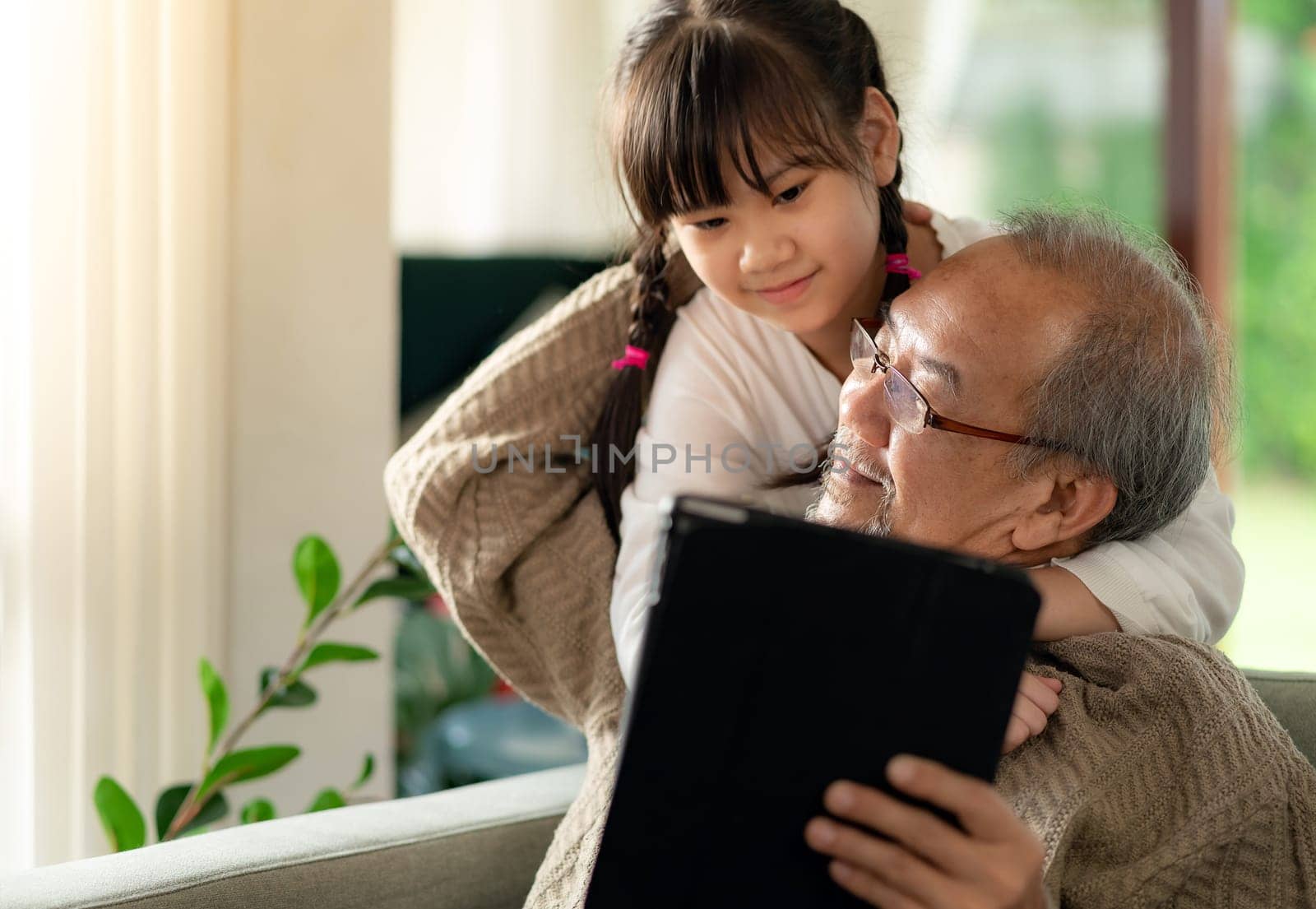 Happy retirement elderly man sitting on sofa at living room with granddaughter using digital tablet together. Multigenerational family with technology concept.