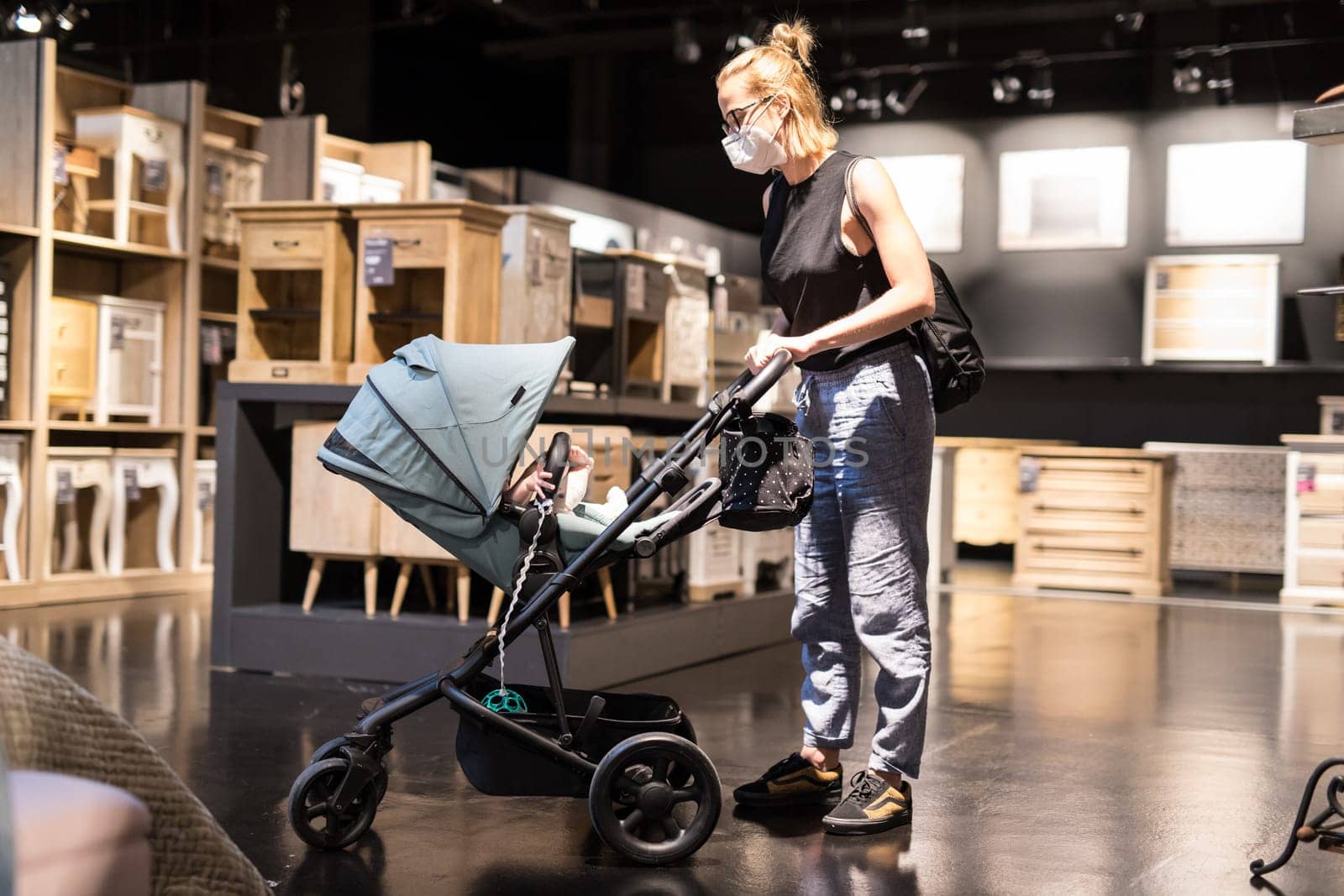Young mom with newborn in stroller shopping at retail furniture and home accessories store wearing protective medical face mask to prevent spreading of corona virus. New normal during covid epidemic.