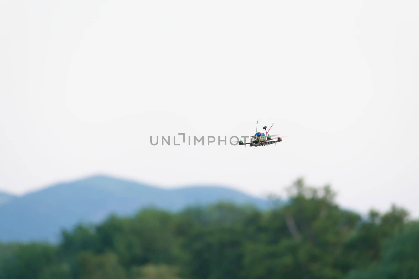 Colorful propellers lift this racing FPV quad copter drone through the air. Plain white sky background with blurred hills and trees beneath