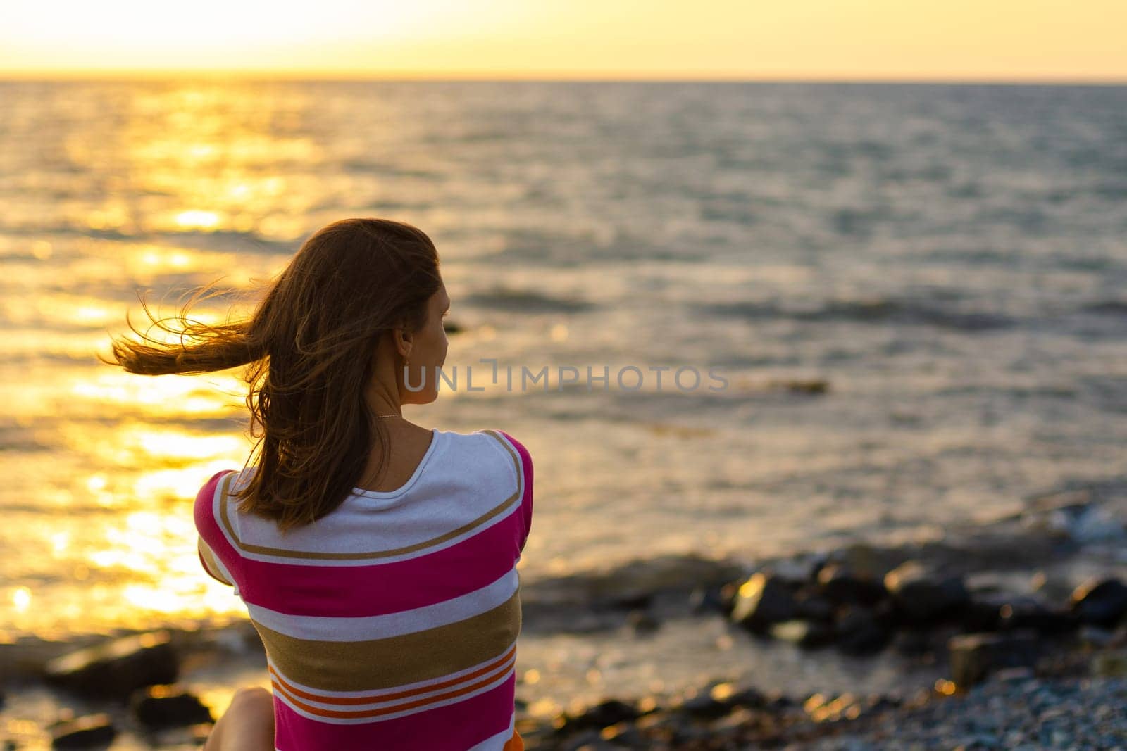 A girl sits in the evening at sunset on the seashore, view from the back