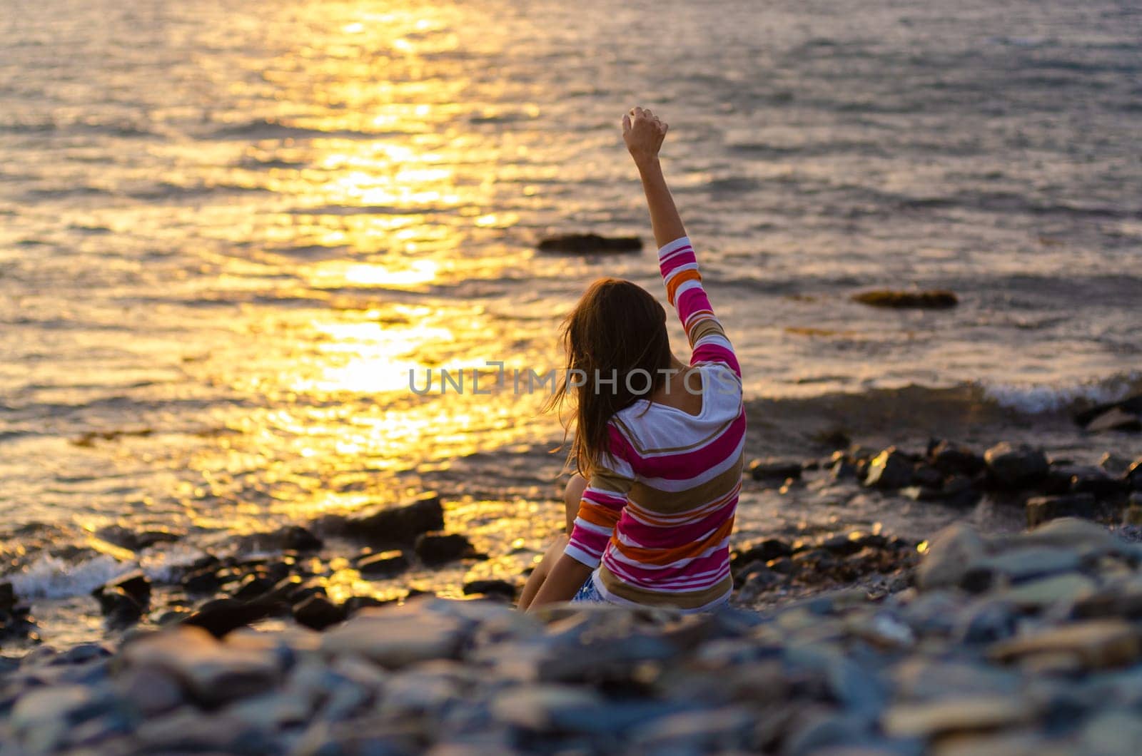 A girl sits on the seashore and throws stones into the water, raised her hands, evening, sunset