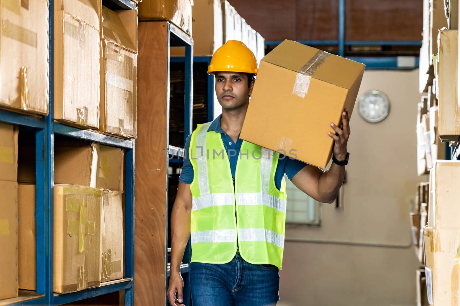 Portrait of Indian asian warehouse worker hold cardboard box packaging on his shoulder in warehouse distribution center environment. Using in business warehouse and logistic concept.