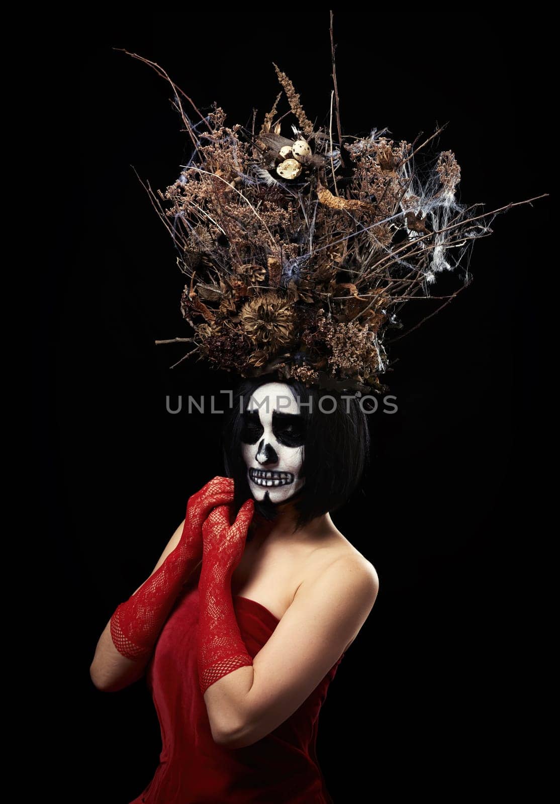 woman of Caucasian appearance with skeleton make-up stands in a red velor dress with a large crown of dry branches, black background