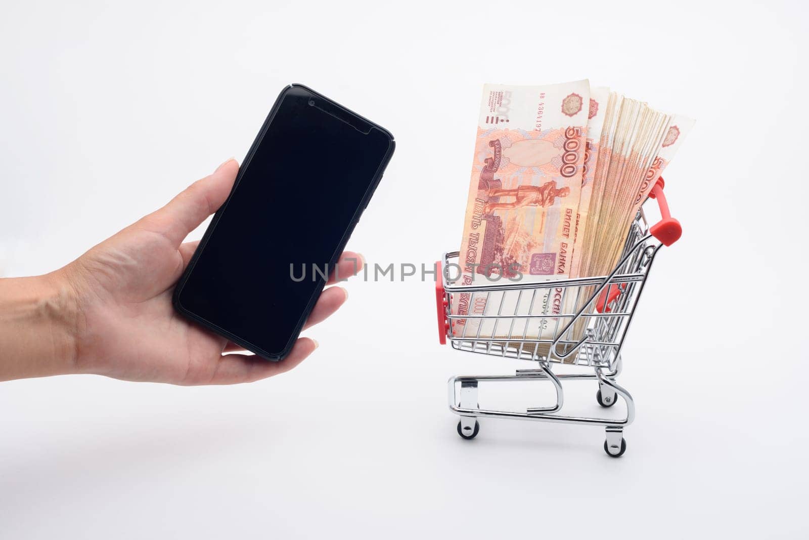 A hand holds a smartphone, next to a grocery cart lies a pile of five-thousandth bills