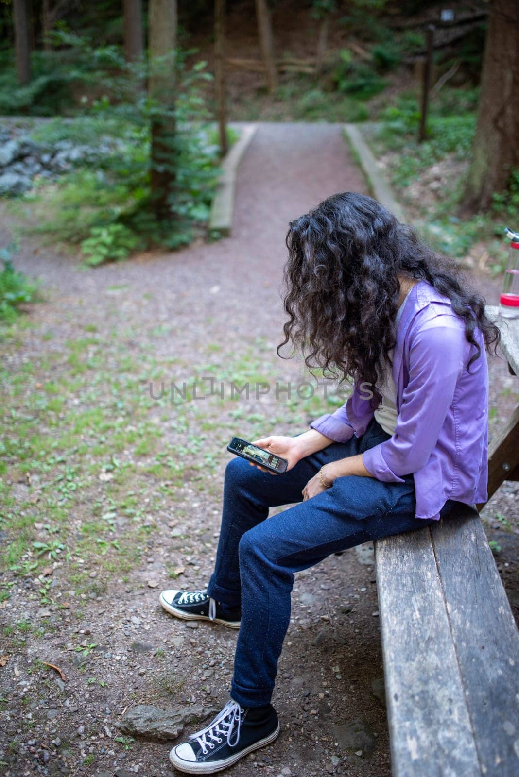 A young caucasian man with long hair and a purple shirt sits slumped at a picnic table along a hiking trail looking at his phone.