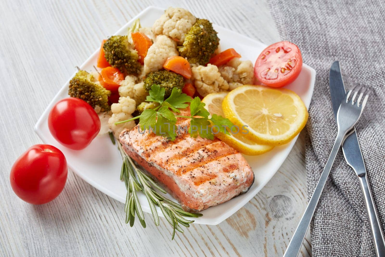 Grilled salmon fillet  garnished with vegetables, lemon, tomato, herbs lying on plate on wooden background. Top view.Healthy food. Keto diet.