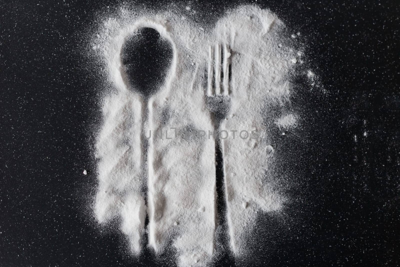 White sugar on black background with silhouette of spoon and fork.