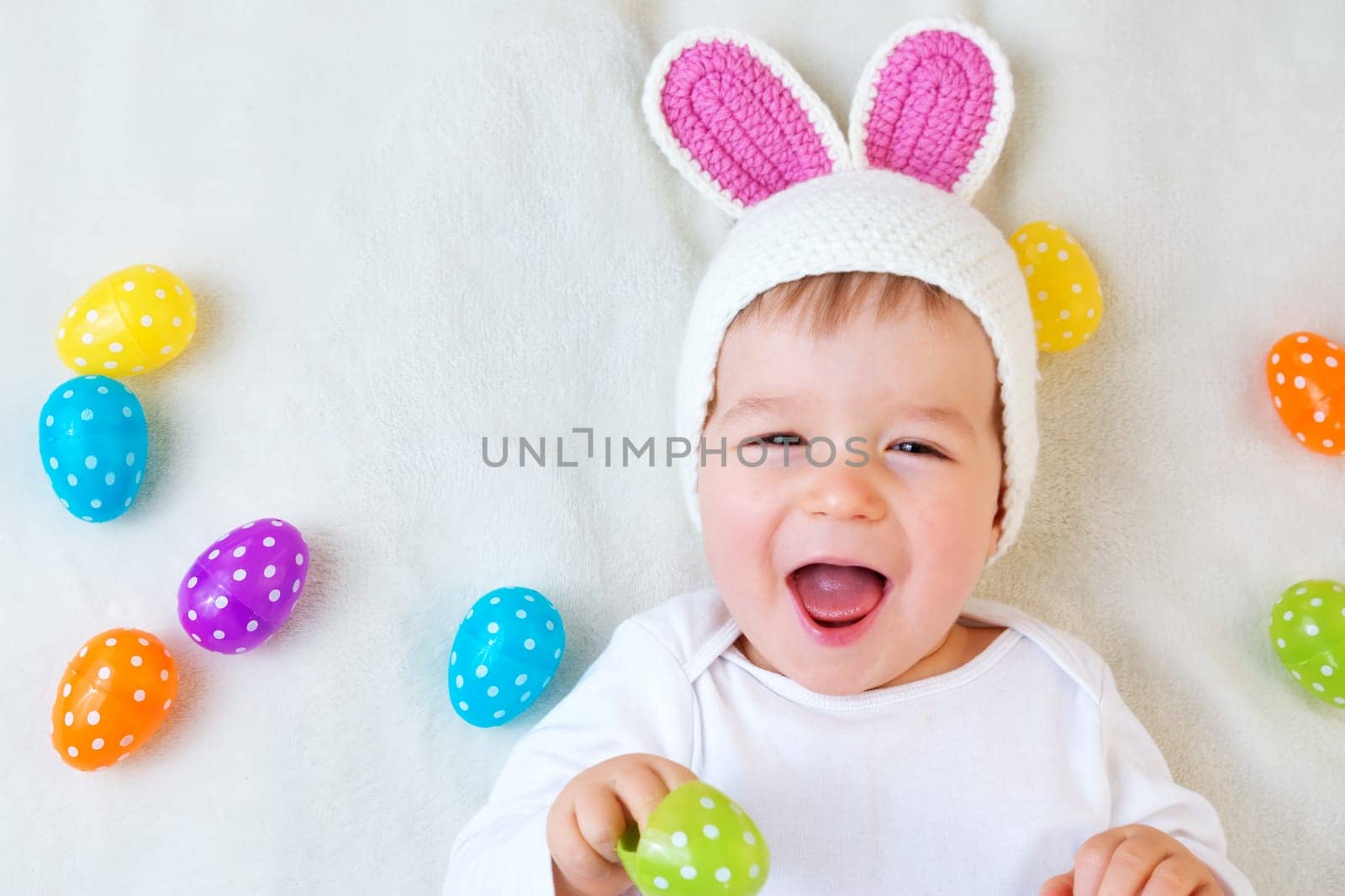 Baby boy in bunny hat lying on green blanket with multicolored easter eggs