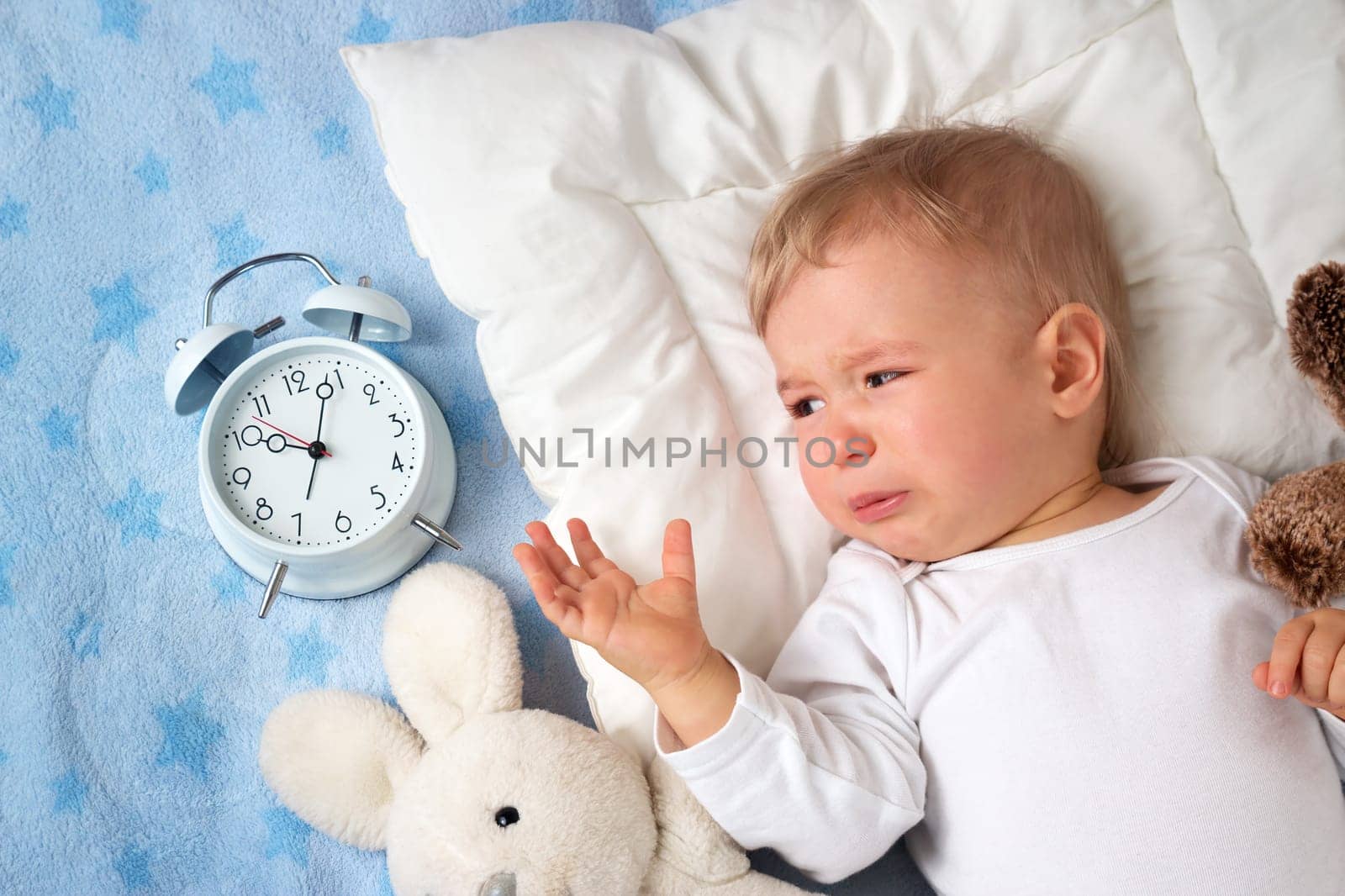 One year old baby lying in bed with alarm clock and crying