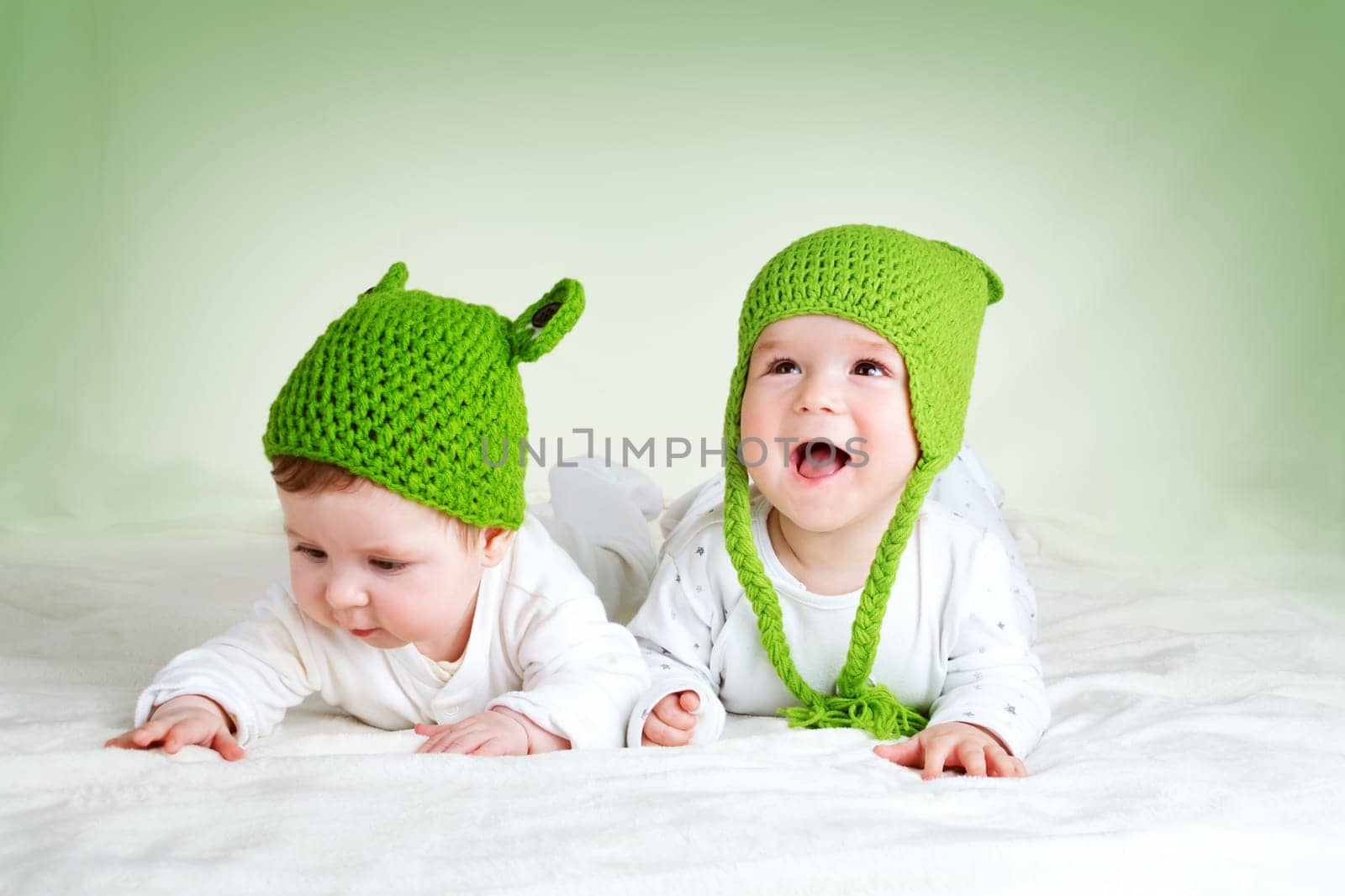 two cute six month old babies lying in frog hats on soft blanket
