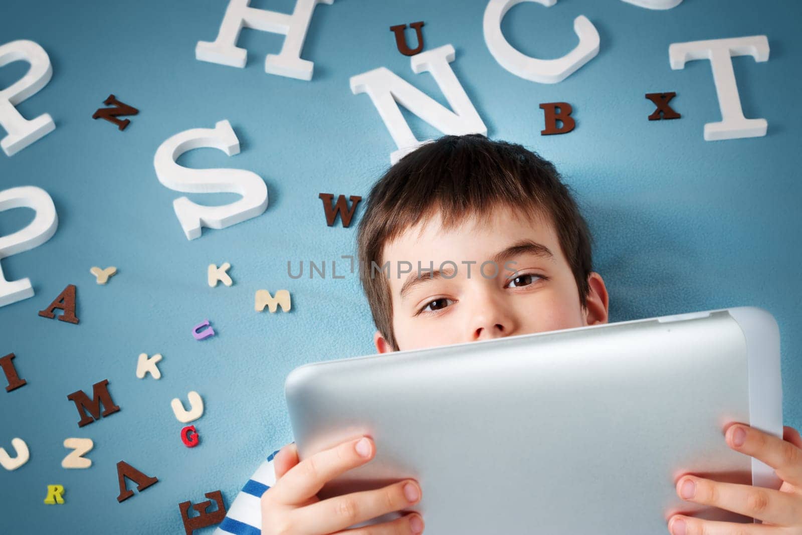 seven years old child lying with tablet and letters on blue background