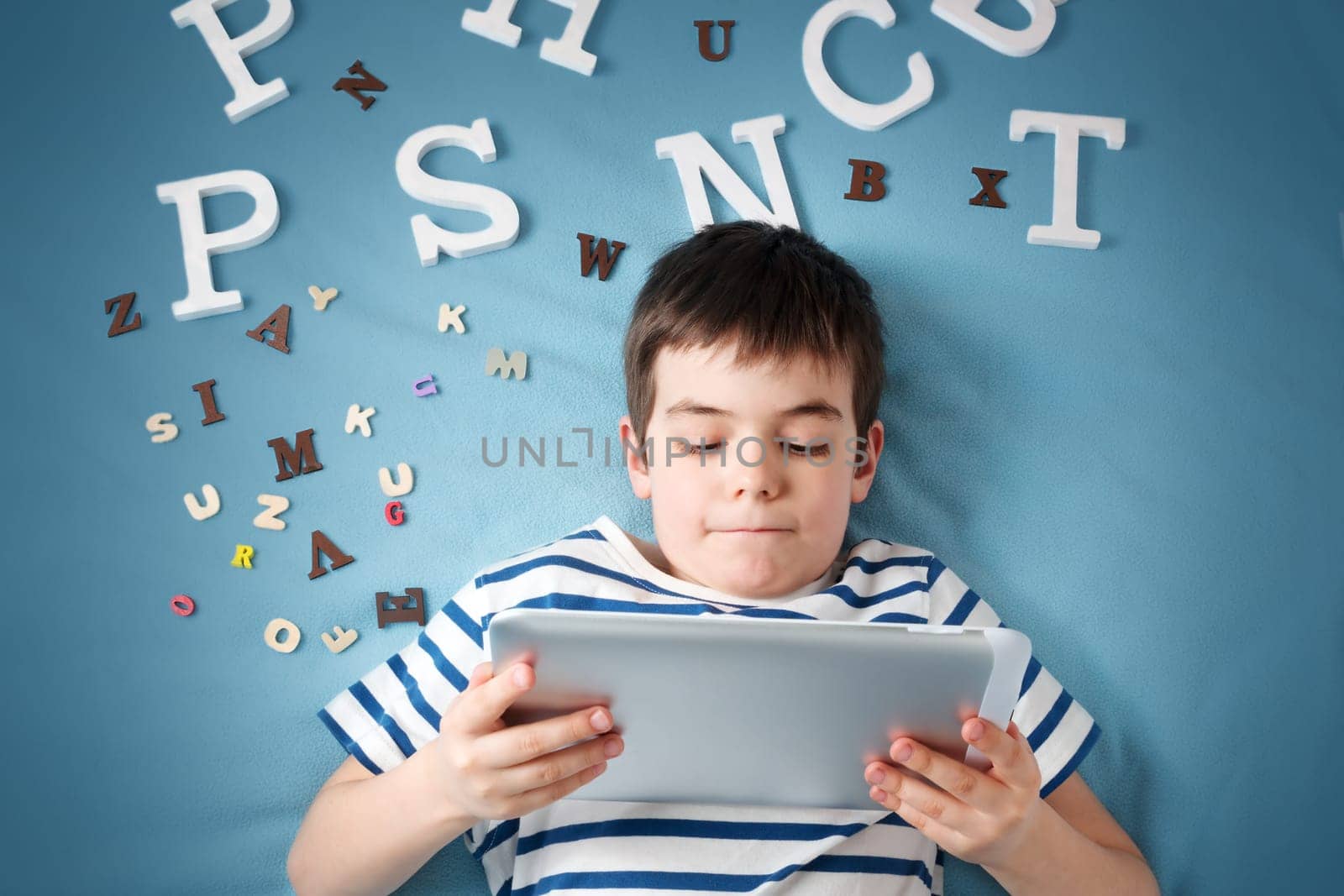 seven years old child lying with tablet and letters on blue background